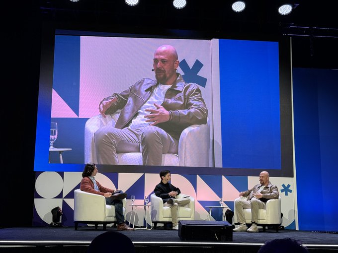 A photograph of a panel discussion on stage with three seated speakers, while a large screen behind them displays a close-up of a bald man in a green jacket speaking.