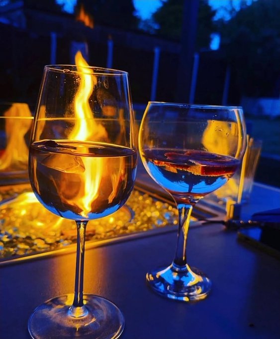 Two wine glasses sit on a table at dusk, one with flames dancing inside and the other holding a red drink, with a fire pit glowing in the background.