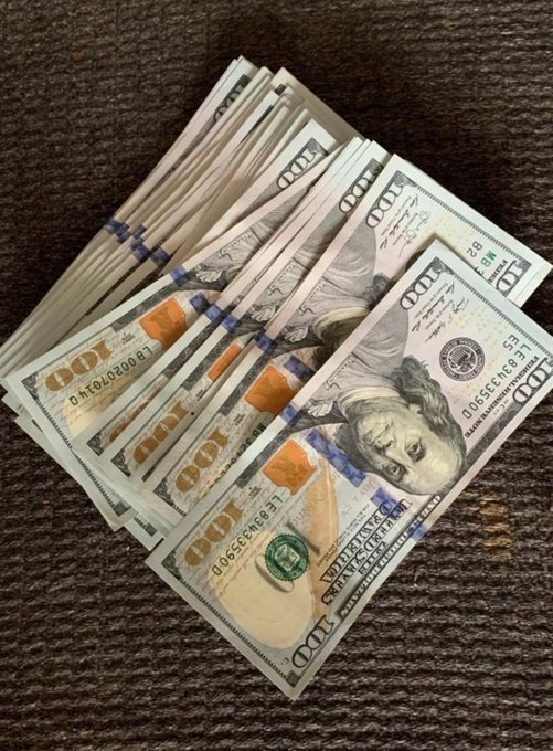 A fan of crisp $100 bills lies fanned out on a dark wood surface, directly illustrating the caption's promise of monetary giveaways to lucky recipients who like the post.