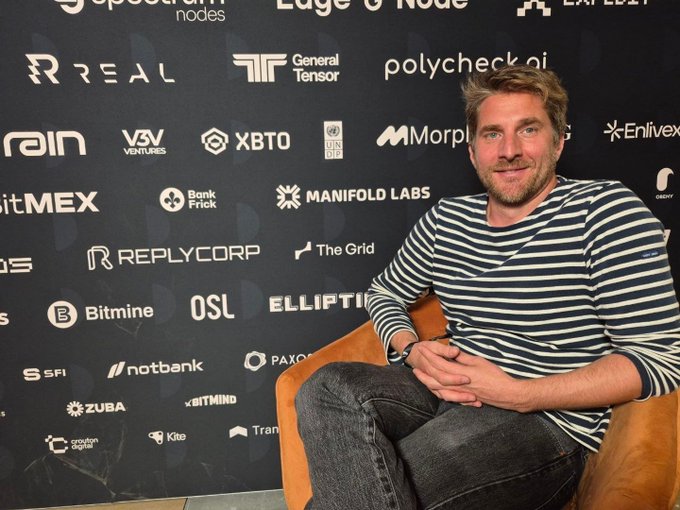 A man in a striped shirt sits in a tan chair at what appears to be a crypto/tech industry event, posing in front of a sponsor backdrop featuring logos like "Ledger," "XBTO," "BitMEX," "Polygon," "Manifold Labs," and "REPLYCORP." The image accompanies coverage of Ledger's response to ZachXBT's criticism, showcasing the company's presence in the Web3/AI space as they push their new AI-agent portfolio management feature that still requires hardware device approval for sensitive actions.