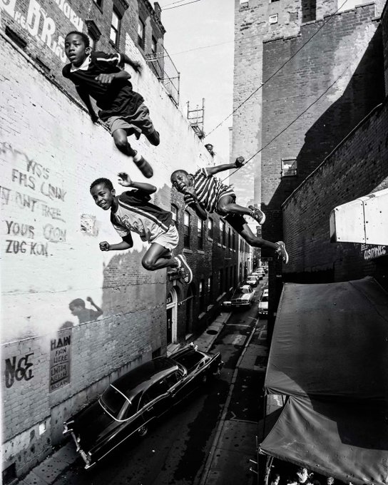 A black-and-white street photograph captures four young boys leaping mid-air between brick buildings in an alleyway, with a vintage car parked below and graffiti visible on the walls.