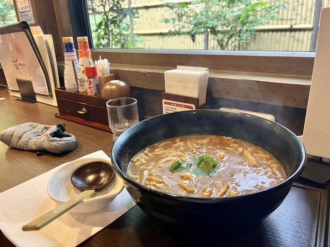 A photograph of a bowl of Japanese curry udon or soup with garnishes, served at a restaurant counter with condiment bottles, a window showing bamboo outside, and a small pouch nearby.