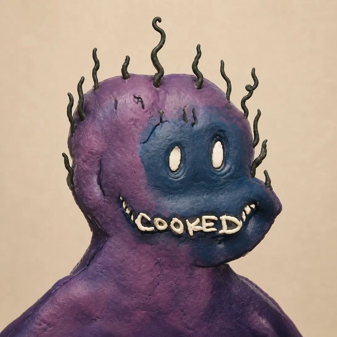 A clay sculpture of a creepy, purple and blue creature with spiky black protrusions on its head, glowing white eyes, and the word 'COOKED' formed by its teeth.