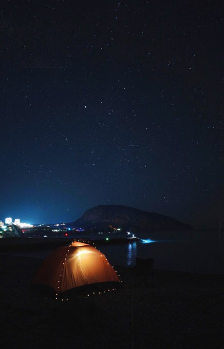 A glowing orange tent is pitched on a sandy beach at night, with a star-filled sky overhead and distant coastal lights illuminating the shoreline.