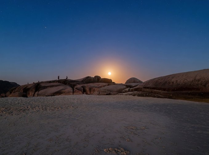 A moonlit coastal landscape with two silhouetted figures standing on smooth granite rock formations under a starry sky, with the glowing full moon rising low on the horizon above the water. The ethereal nighttime scene, titled "Midnight Glow," perfectly complements the post's invitation to collect this edition, capturing the radiant lunar illumination that gives the artwork its name.