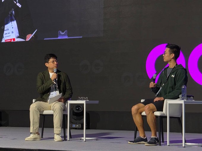 Two men are seated on a stage having a discussion, with one holding a microphone and papers while the other holds a microphone, set against a backdrop featuring purple circular logos. A water bottle and a book rest on small tables beside them, suggesting a casual fireside chat or interview setting—fitting the simple sentiment of "thanks" for a speaking engagement or panel appearance.