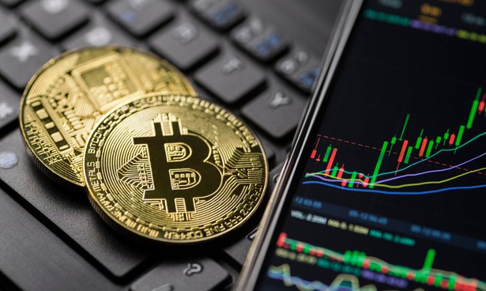 Two physical Bitcoin coins rest on a black laptop keyboard beside a smartphone displaying a candlestick price chart, visually representing the $BTC market analysis and the critical $81,822 weekly resistance level that bulls must break to confirm a broader uptrend.