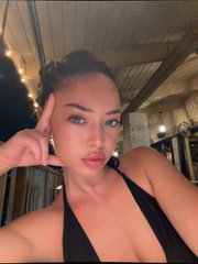 A selfie of a woman with dark hair pulled back, posing with her hand on her head and a sultry expression, wearing a black top. The caption pairs with the post about emotional unavailability, using the detached, all-in-on-myself look to humorously mirror the cold, disengaged vibe of a crypto trader ignoring feelings to focus on charts.