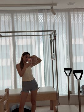 A woman takes a mirror selfie in a bright Pilates studio while holding a resistance ring, capturing her "big family" gym welcome to prospective members. The setting, featuring a Pilates reformer and dumbbells, visually reinforces the post's invitation to join her fitness community.