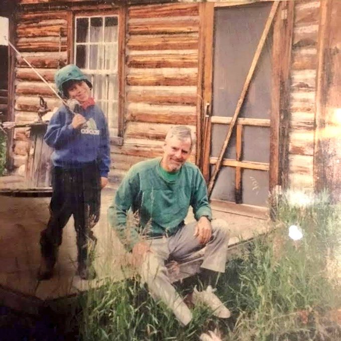 The caption should be:

A young boy in a blue Adidas hoodie and green cap stands next to his smiling father in a green sweater outside a rustic log cabin, capturing a tender family moment that contrasts with the post's joke about the boy refusing to help sell Bitcoin.
