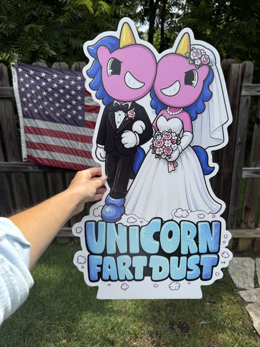 A hand holds up a cartoon wedding yard sign featuring two pink unicorns dressed as bride and groom, with the text "UNICORN FART DUST" displayed below, set against an American flag backdrop—visually representing the whimsical $UFD memecoin community and the playful love story described in the post.
