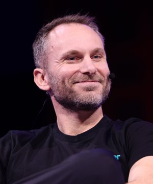 A close-up of a smiling man with short grey hair and a beard, wearing a black t-shirt and a headset microphone.