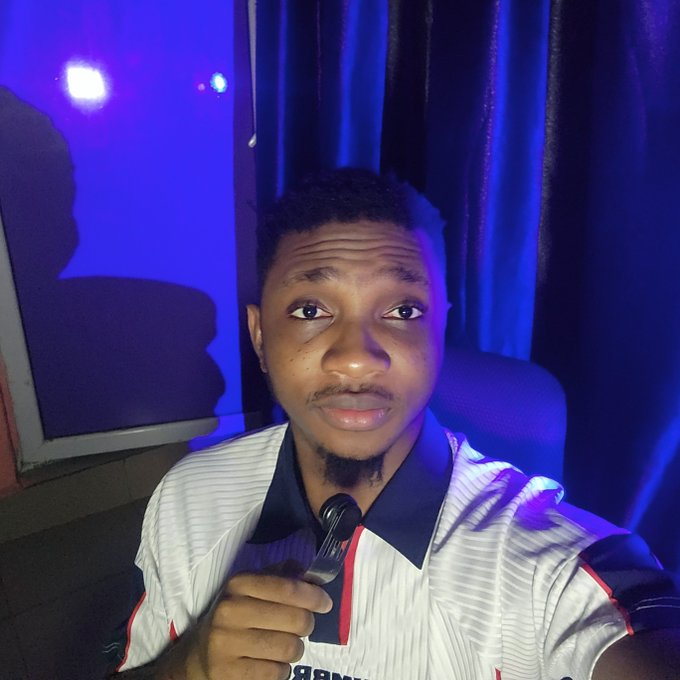 A young man with a goatee wearing a white and dark blue striped polo shirt holds a small microphone, sitting in a room illuminated by dramatic blue lighting.
