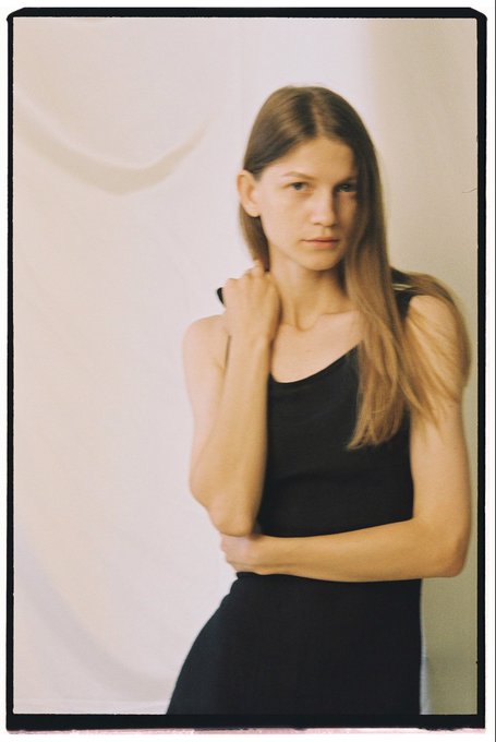 A portrait photograph of a young woman with long light brown hair, wearing a black sleeveless top, posing against a white backdrop with her arms crossed.