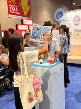 Fans browse merchandise and photocards at a TWICE-related booth or convention, with a 'FREE Giveaways' sign visible and a young woman looking at album photocards while wearing a tote bag with plush keychains.