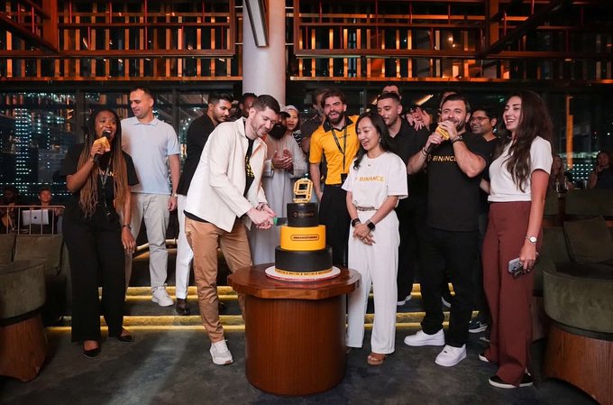 A group of Binance team members gather around an elegant three-tiered cake decorated with a golden "9" topper, celebrating the company's 9th anniversary in a high-rise venue with city views. The cake's central "9" perfectly captures the milestone reflected in the "#binanceturns9" celebration.