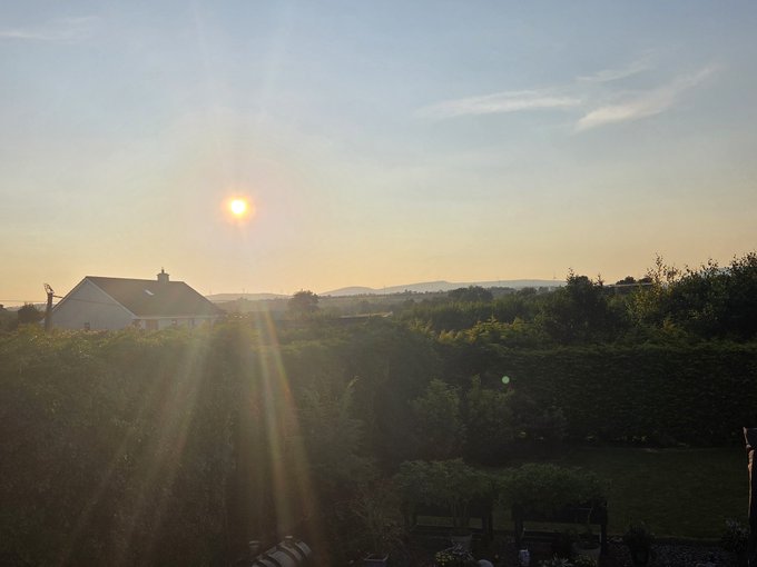 A golden sun sets over a rural landscape in the West of Ireland, casting long rays across green hedgerows and a white cottage, illuminating the scenic countryside that ties together themes of Bitcoin, hope, and freedom.