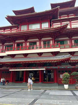 A multi-tiered building with traditional Chinese architectural elements, featuring red columns, upturned eaves, and gray tiled roofs, with a person in a white shirt standing on the paved plaza in front.