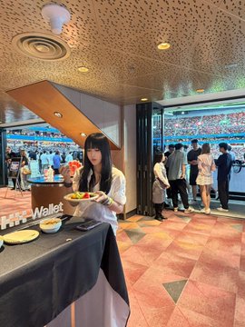 A young woman poses with a plate of food at an OKX-sponsored lounge overlooking a soccer pitch, where fans and players are visible on the field behind her—matching the post's theme of enjoying a live soccer match as an OKX guest.