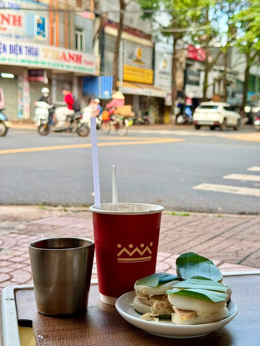A street-side view of a red paper cup with a crown logo, a metal tumbler, and a plate of small food items on a table, with a Vietnamese street scene in the background.