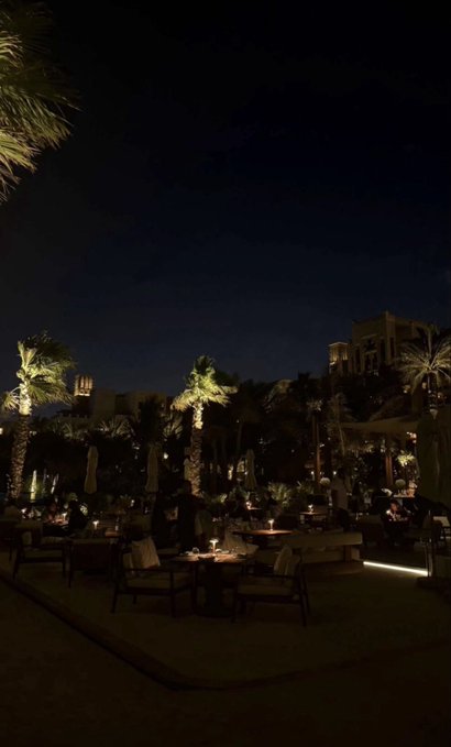 A dimly lit outdoor lounge area features palm trees, small candle-lit tables, and warm lights against a dark night sky.