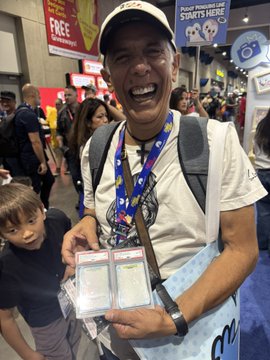A smiling man wearing a cap, lanyard, and backpack holds what appears to be collectible card packs at what looks like a comic or collectibles convention.