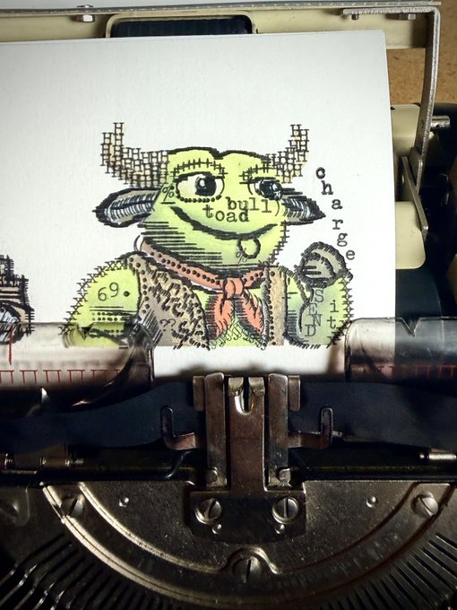 A close-up of a vintage typewriter with paper showing a stamped, collage-style illustration of a horned character and the words 'bull toad' and 'charge it'.