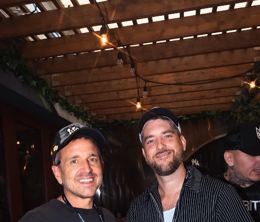 Two smiling men pose together at what appears to be an outdoor evening venue, one wearing a dark baseball cap and the other a white cap with sunglasses perched on top.