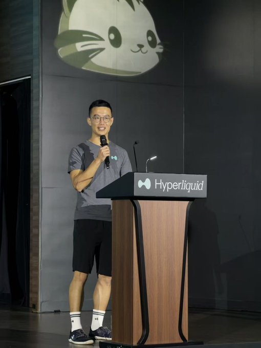 A young man with glasses stands at a wooden lectern holding a microphone, with "Hyperliquid" displayed on the podium and a cartoon cat mascot projected on the wall behind him, matching the post's mention of Jeff excitedly shouting "Hyperliquid!"