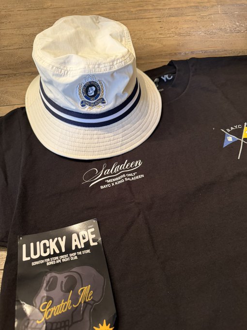 A white bucket hat with an emblem sits atop a black t-shirt featuring 'Saladeen' and 'BAYC x KONG BALADEEN' text alongside a 'Lucky Ape Scratch Me' packet.