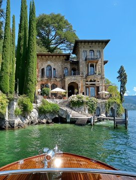 A view from a wooden boat approaching a luxurious stone villa with tall cypress trees, overlooking clear turquoise water under a blue sky.