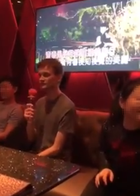 A dimly lit karaoke room where a young man holds a pink microphone in front of a TV screen displaying Chinese karaoke lyrics.