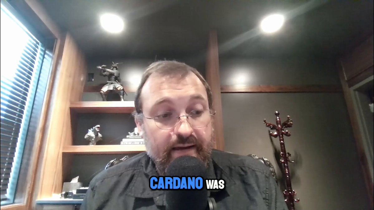 The image shows Charles Hoskinson talking into a microphone during an online broadcast or video call. He appears to be wearing glasses and has a beard. The background of the image features a bookshelf with various items, including a statue on top. The caption below the image reads "If we're real, we can survive anything" which is related to Charles Hoskinson's statement about the winners and losers cycle in cryptocurrency markets.