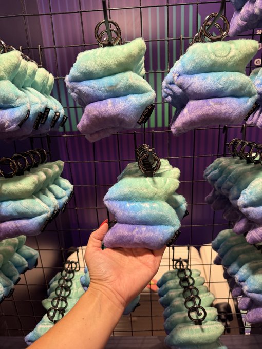 A hand holding a pastel blue and purple tie-dye plush keychain, displayed in front of a wire grid rack filled with identical plush items in a retail setting.