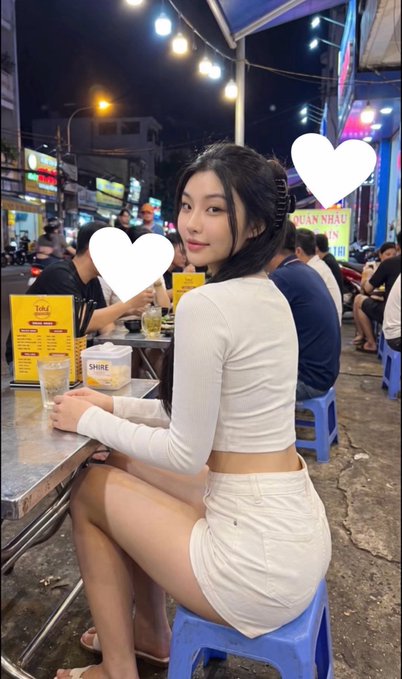 A young woman in a white long-sleeve crop top and white skirt sitting on a blue plastic stool at an outdoor street food stall at night, with white heart emojis overlaid on the image.