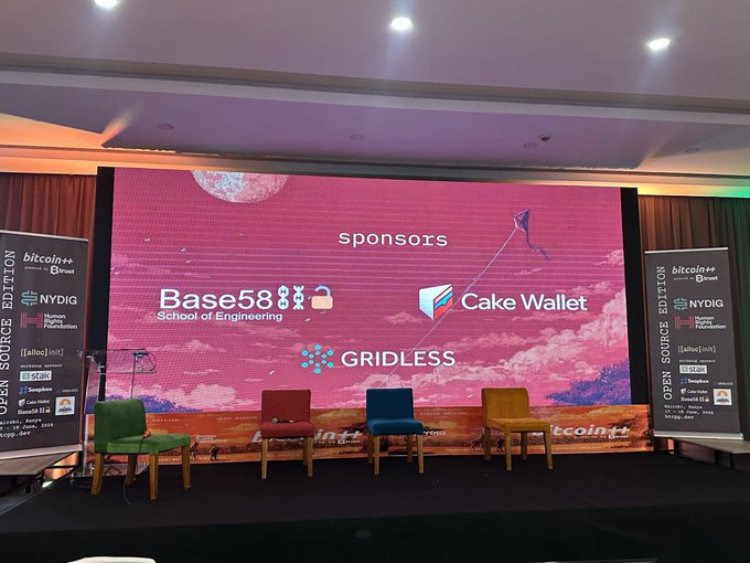 The image shows an event stage with a large screen displaying a pink background, featuring logos for Base58, Cake Wallet, and Gridless. There are three chairs arranged around the stage, each adorned with a different colored cushion. The stage also has a microphone on its left side. In front of the stage, there is a banner that reads "Amazing first day at @btcplusplus Nairobi!".