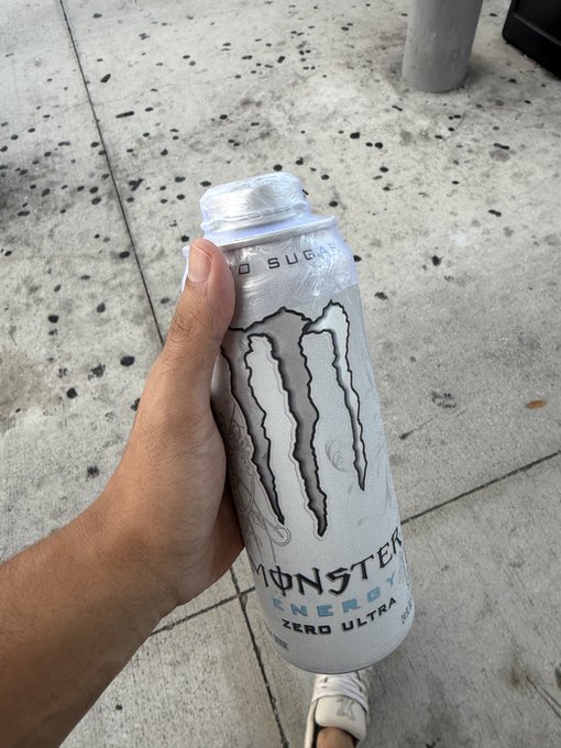 A hand holding a white Monster Energy Zero Ultra sugar-free can on a concrete sidewalk, with the person's white sneaker visible at the bottom of the frame.
