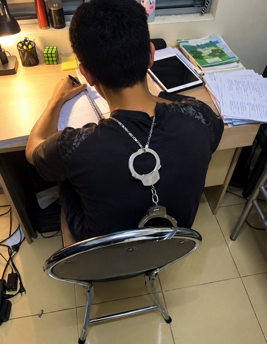 A young man sits at a desk with handcuffs around his wrists and a chain connecting his wrists to the back of his chair; he is writing in a notebook with various school supplies nearby.