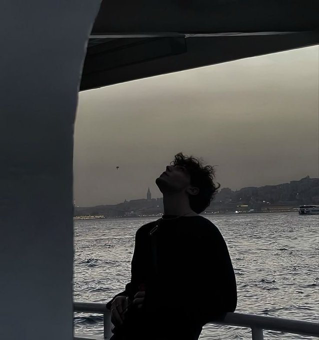 Moody silhouette photograph of a person in a dark hoodie leaning back with their face tilted up, standing by a railing overlooking water at dusk with a distant city skyline.