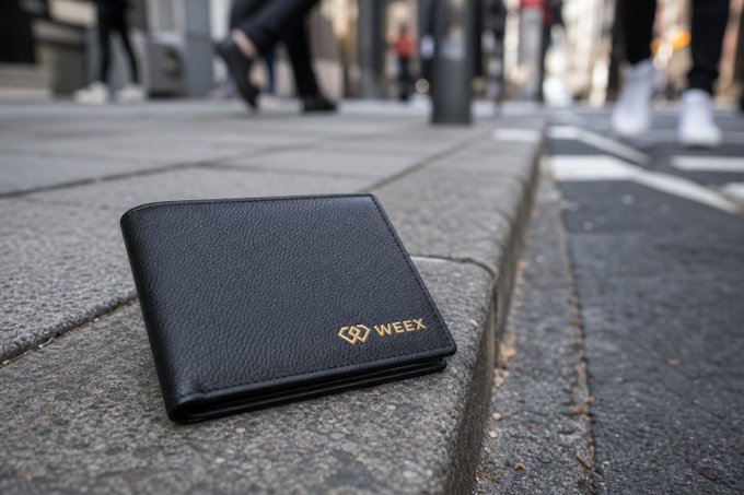 The image features a black wallet laying on its side, placed on a sidewalk or street curb. The wallet has gold lettering that reads "weex" along with other text. There are also people walking by in the background, indicating an urban setting.
