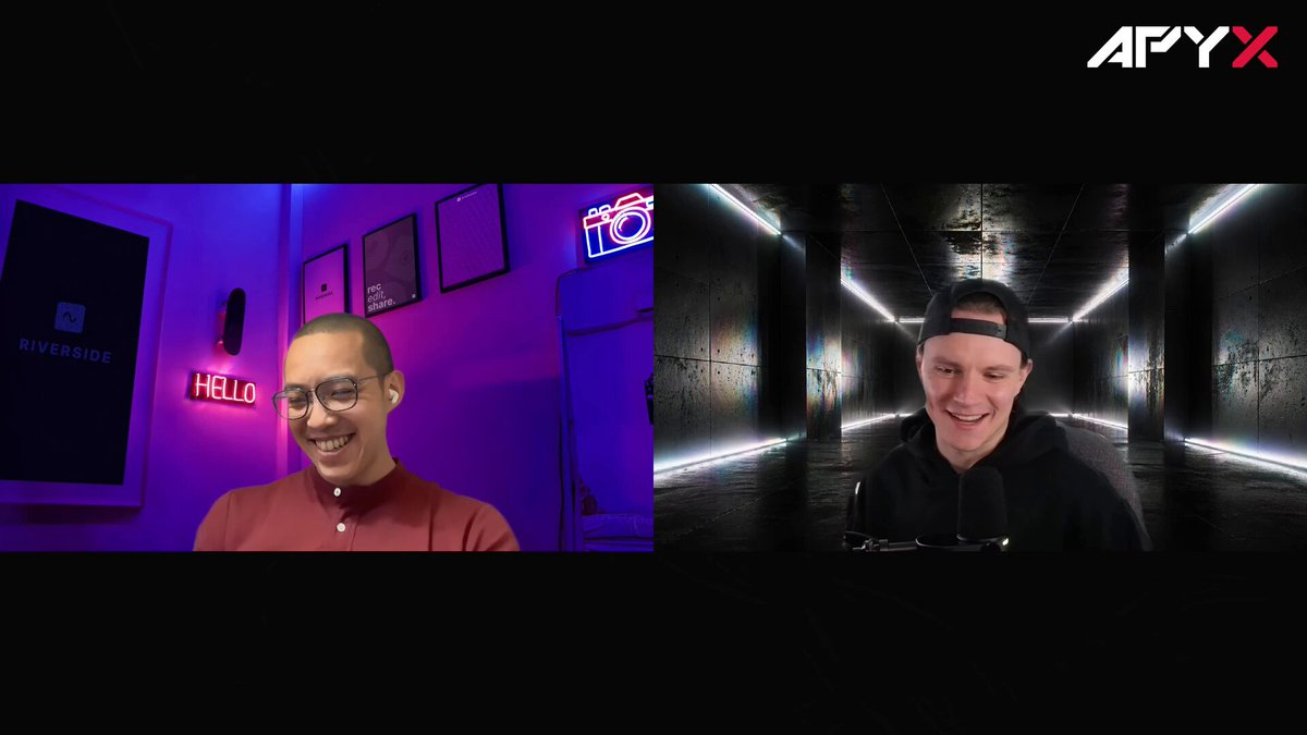 The image shows two men sitting next to each other at a computer desk with their faces displayed on a video call screen. They are both smiling for the camera while engaged in conversation. The background of the image features a black wall, which contrasts with the colorful neon lights that illuminate the room. There is also a refrigerator visible in the scene.