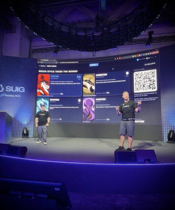 Two men present an interactive NFT arena game on a stage, with a large screen displaying categories like 'GIPPING TOAD', 'UNDERTOW', 'SKINNER', and 'ANACODON' alongside a QR code for voting.
