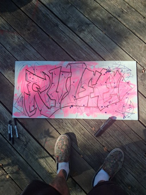 A first-person view looking down at camo-patterned sneakers standing on a wooden deck next to a graffiti artwork spelling 'RICH' in pink spray paint, with two markers and a paint bottle nearby.