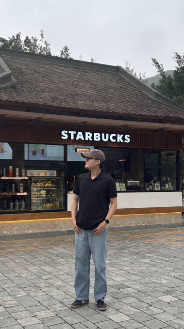 A man wearing a cap, black polo shirt, and light blue jeans stands in front of a Starbucks coffee shop.