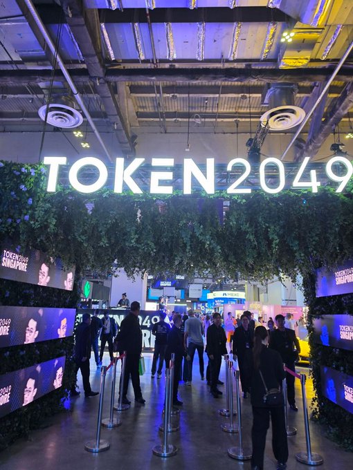 A large illuminated 'TOKEN2049' sign hangs above an entrance to what appears to be a Singapore conference venue, with attendees walking through a leaf-decorated archway.
