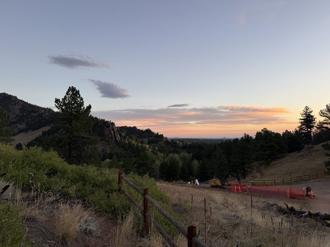A landscape photograph taken at sunset or sunrise showing a rural scene with a wooden split-rail fence, rolling hills covered in trees, and orange safety fencing with equipment in the distance under a sky with scattered clouds.