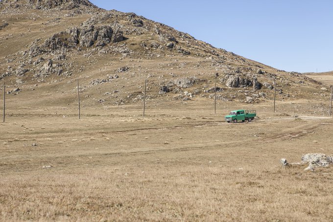 A green utility truck driving along a dirt road in a vast dry, rocky, hilly landscape with sparse vegetation under a clear sky.