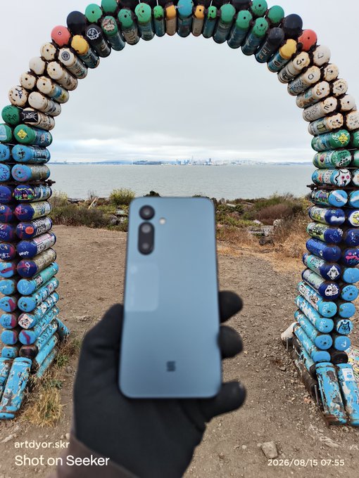 A gloved hand holding a light blue smartphone with three camera lenses, framed by an archway made of spray-painted bottles, looking out over water with a city skyline in the distance; the image is marked 'artdvor.skr Shot on Seeker' dated 2026/08/15 07:55.