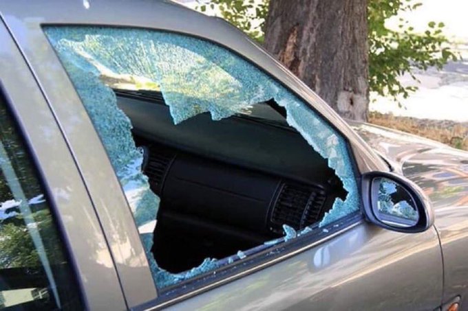 A silver car with a completely shattered driver's side window, with broken glass still clinging to the frame.