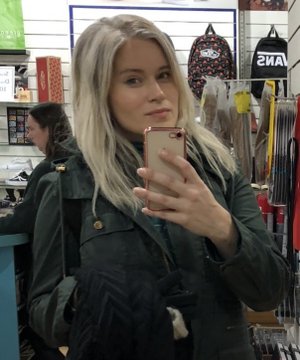 A woman with long blonde hair taking a mirror selfie with a rose-gold smartphone while wearing a dark denim jacket, with another shopper visible in the background of what appears to be a retail store.